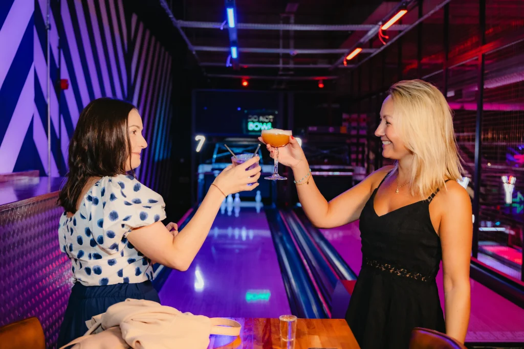 Two women toasting their drinks in front of the duck pin bowling lanes at Roxy